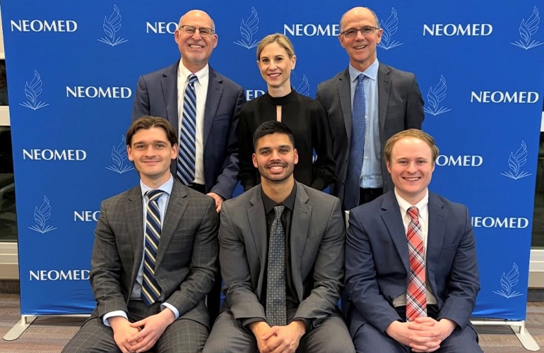 Six people in business attire pose in front of a NEOMED backdrop