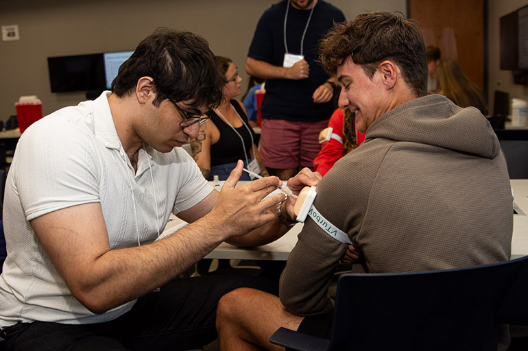 A young man in a white polo shirt practices injecting a needle into the arm of a young man in an olive-colored hoody