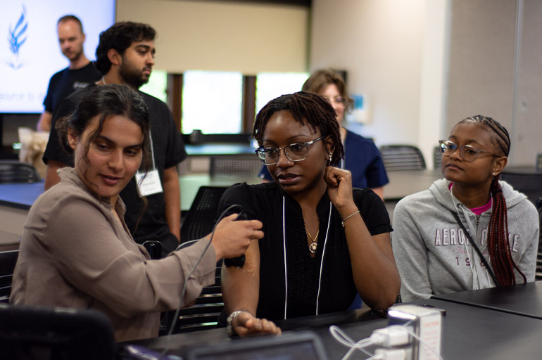 A young woman in a sand-colored shirt practices taking the blood pressure of a young woman in a brown T-shirt as other people look on