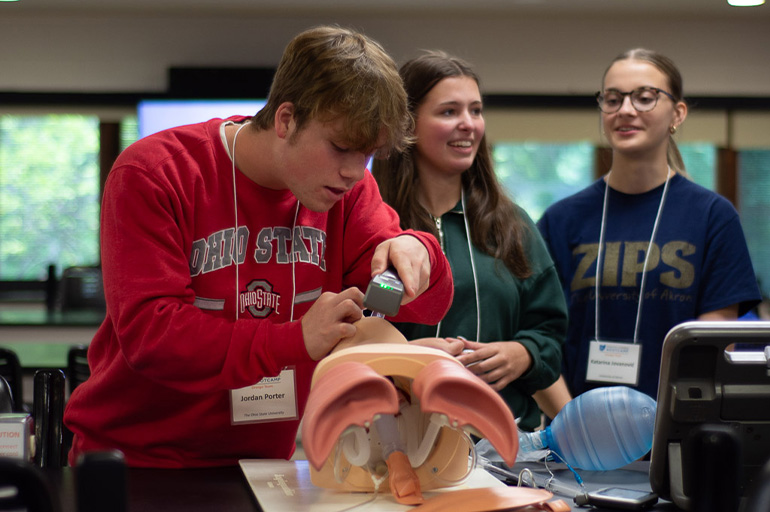 A young man in a red sweatshirt practices intubation on a medical simulation manikin as two young women look on
