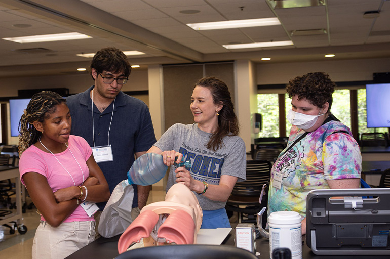 A young woman in a NEOMED T-shirt demonstrates intubation on a medical simulation manikin to three onlookers