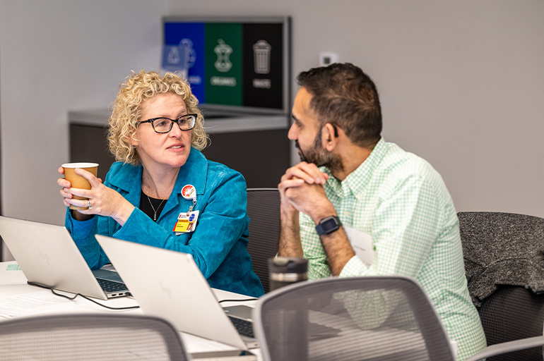 a woman and a man chat while seated at a table, sitting behind laptops