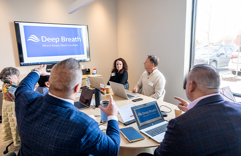 A group of people sit around a conference table in front of a screen that reads "Deep Breath"