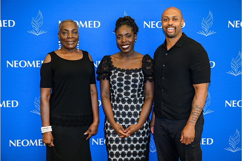 Two women and a man in business casual attire pose in front of a blue NEOMED backdrop