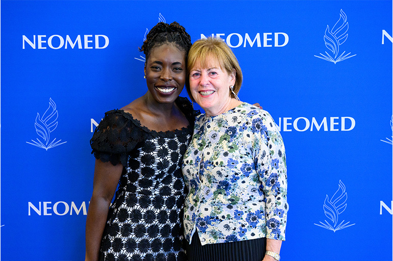 Two smiling women pose in front of a blue NEOMED backdrop