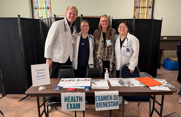 Four students in white coats stand behind a table labeled "Healh Exam" and "Examen de Bienestar"