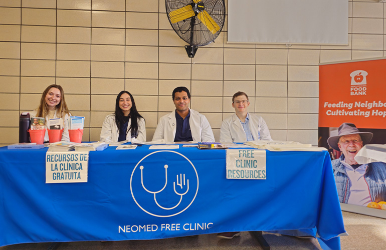 Four students in white coats sit at a table with the NEOMED Free Clinic table cloth draped over it