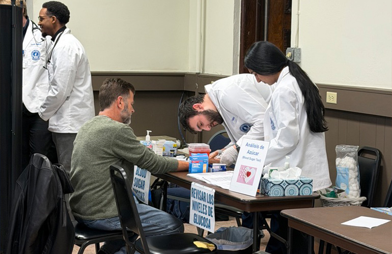 A man in white coat performs a glucose check on a man while a woman supervises