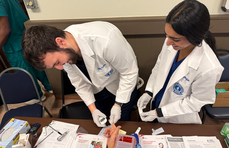 A man in white coat holds a glucose meter up to a patient's hand while a woman in white coat looks on