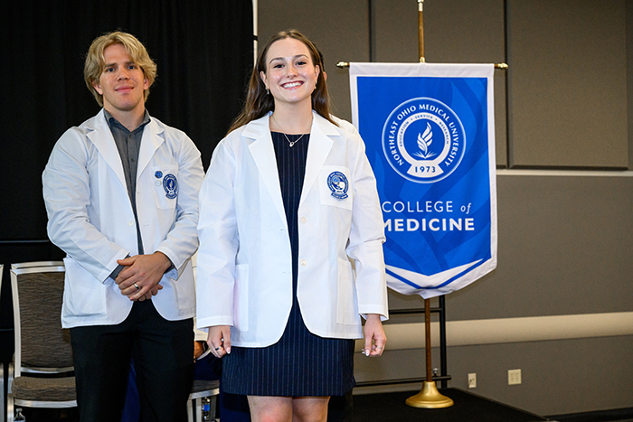 A young man in white coat stands behind a young woman in white coat. A gonfalon for the NEOMED College of Medicine appears in the background.