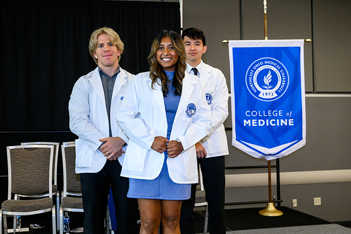 A young woman in white coat stands between and in front of two young man, also in white coats. The gonfalon for the NEOMED College of Medicine appears in the background