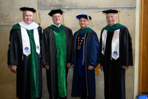 Four men in academic regalia pose in front of a stone wall