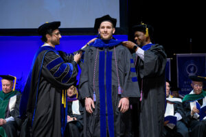 Three men in academic regalia. The man in the center is being hooded with his doctoral hood by the other men