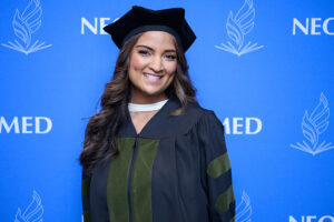 a young woman in academic regalia poses in front of a blue backdrop branded with the NEOMED logo