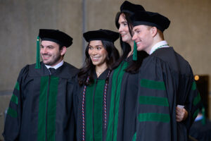 a group of graduating students in academic regalia smile at something outside the frame
