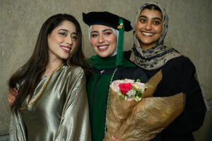 Three young women smile at the camera. The woman in the center wears academic regalia for the NEOMED College of Medicine and holds a bouquet of flowers