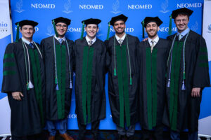 A group of young men in the academic regalia of the NEOMED College of Medicine pose in front of a university backdrop