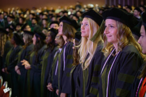 The audience at a commencement ceremony. The focus is on two young women in the foreground who are dressed in academic regalia for the NEOMED College of Pharmacy and are looking upward