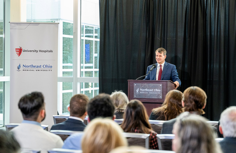 Dr. John T. Langell at podium addresses a group of seated guests while standing next to a UH-NEOMED banner