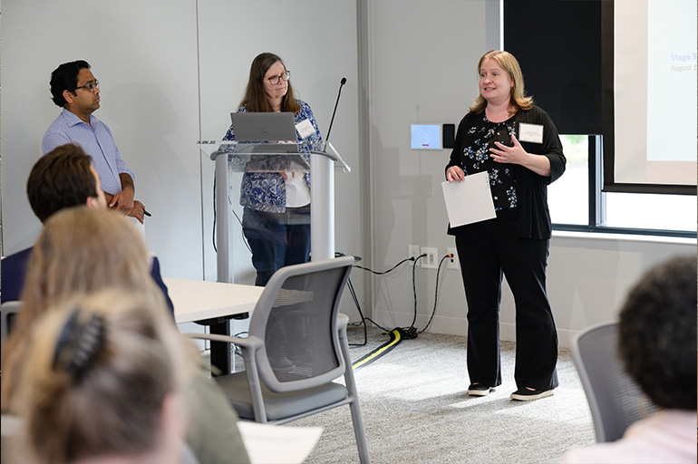Two women and a man stand in front of a classroom. One woman is speaking to the audience and the other two people look on