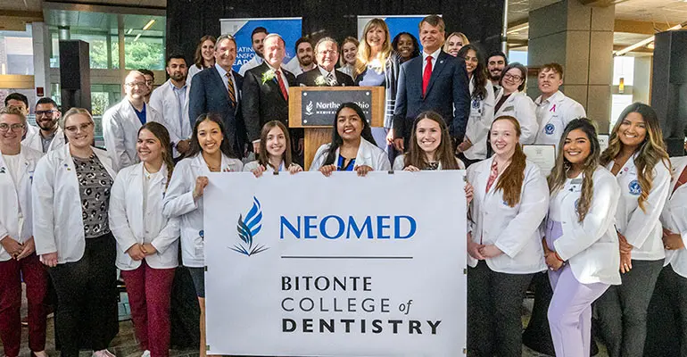 A group of students in white coats stand around a sign that says Bitonte College of Dentistry.
