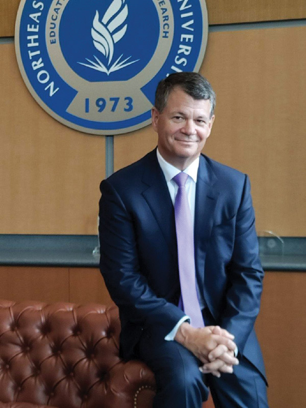 The president of the university, wearing a suit and sitting before the crest of the university on a wall behind him.