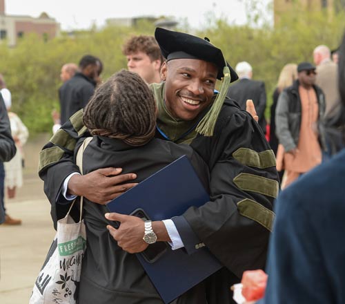 A graduate is all smiles after commencement as he hugs a relative.