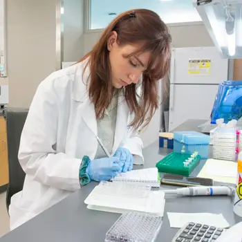 A researcher in a white coat takes notes in a lab.