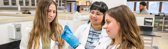 Three pharmacy students in white coats examine a vial in a lab.