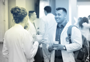Two medical students talk in a bright hallway while enjoying cups of coffee.