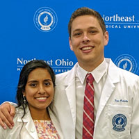 Two medical students in white coats stand before a blue background, smiling.