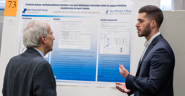 white haired man listens to a young man with beard talk about a scholarly poster.