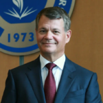 The president of the university, wearing a suit and sitting before the crest of the university on a wall behind him.