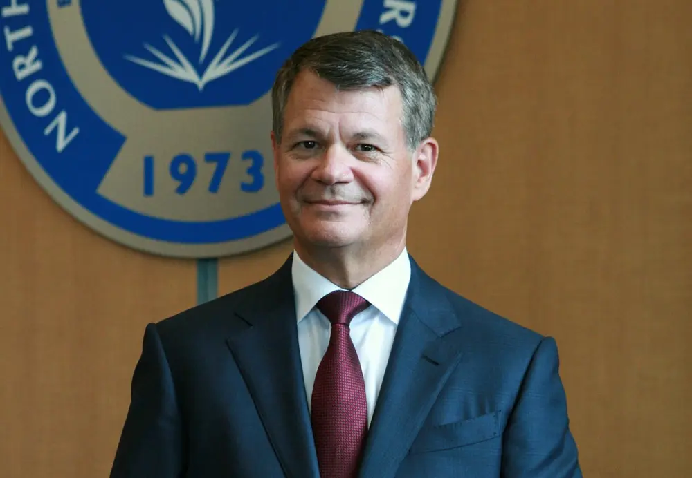 The president of the university, wearing a suit and sitting before the crest of the university on a wall behind him.