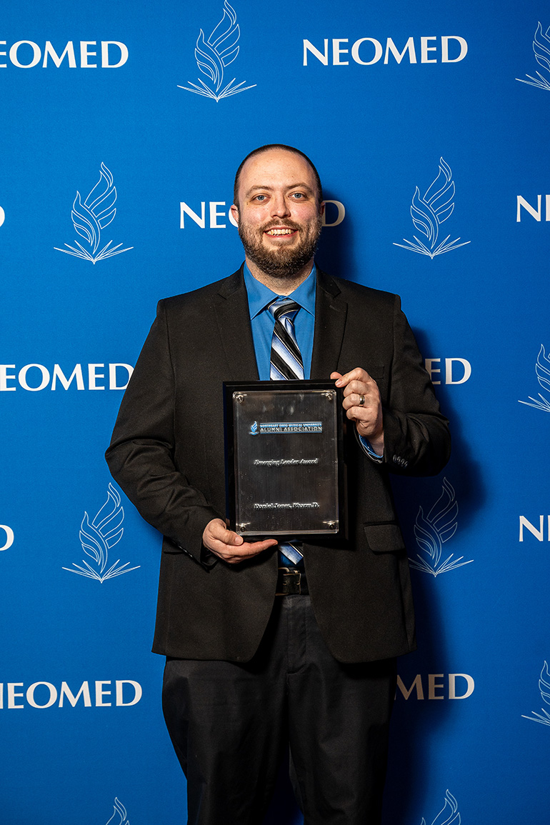 Dan Jones, in suit and tie, poses with an award plaque in front of a blue NEOMED backdrop