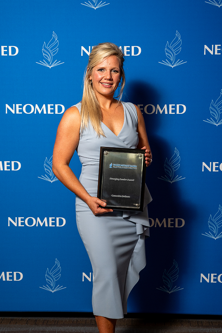 Casondra Seibert, in a light blue dress, poses with an award plaque in front of a blue NEOMED backdrop