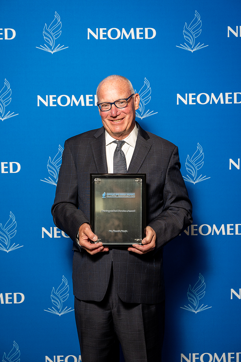 Frank Beck, in suit and tie, poses with an award plaque in front of a blue NEOMED backdrop