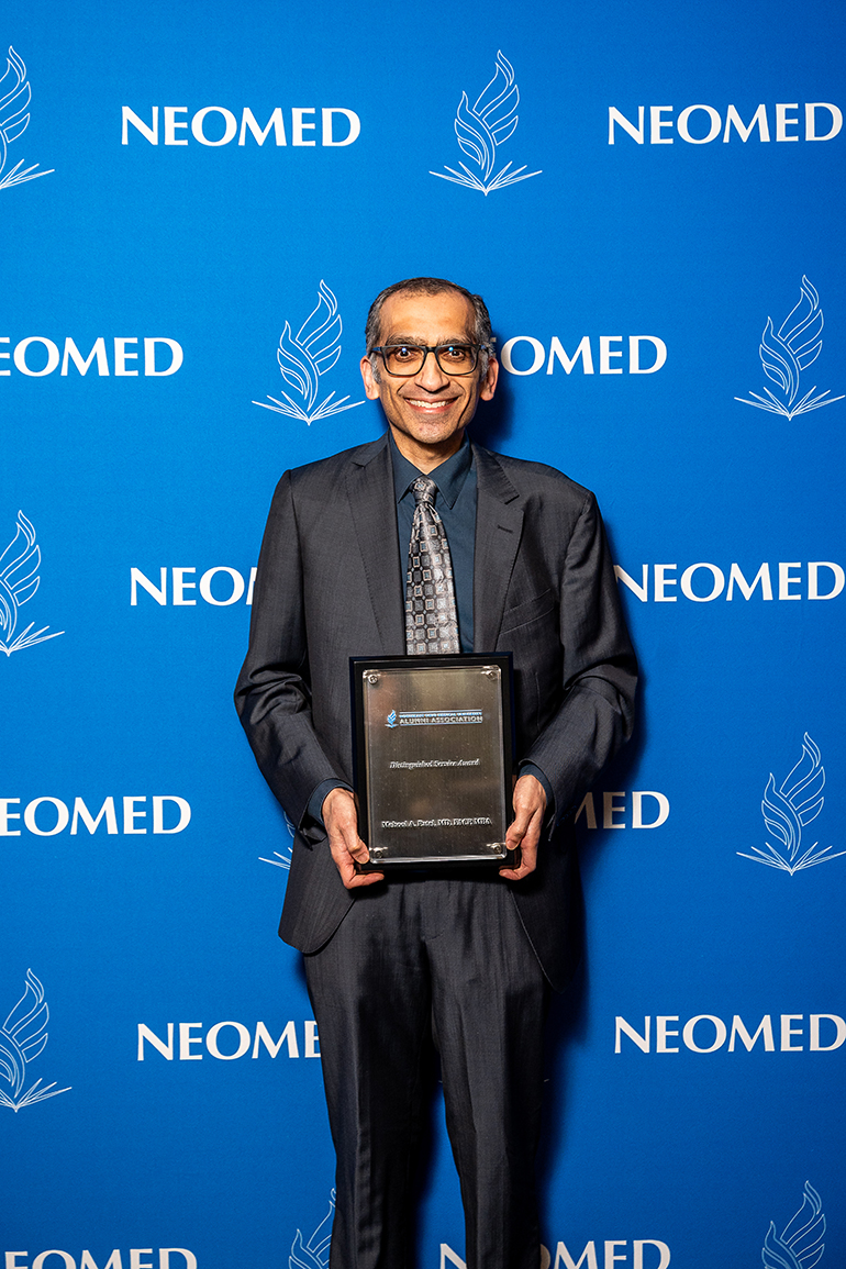 Mehool Patel, in suit and tie, poses with an award plaque in front of a blue NEOMED backdrop