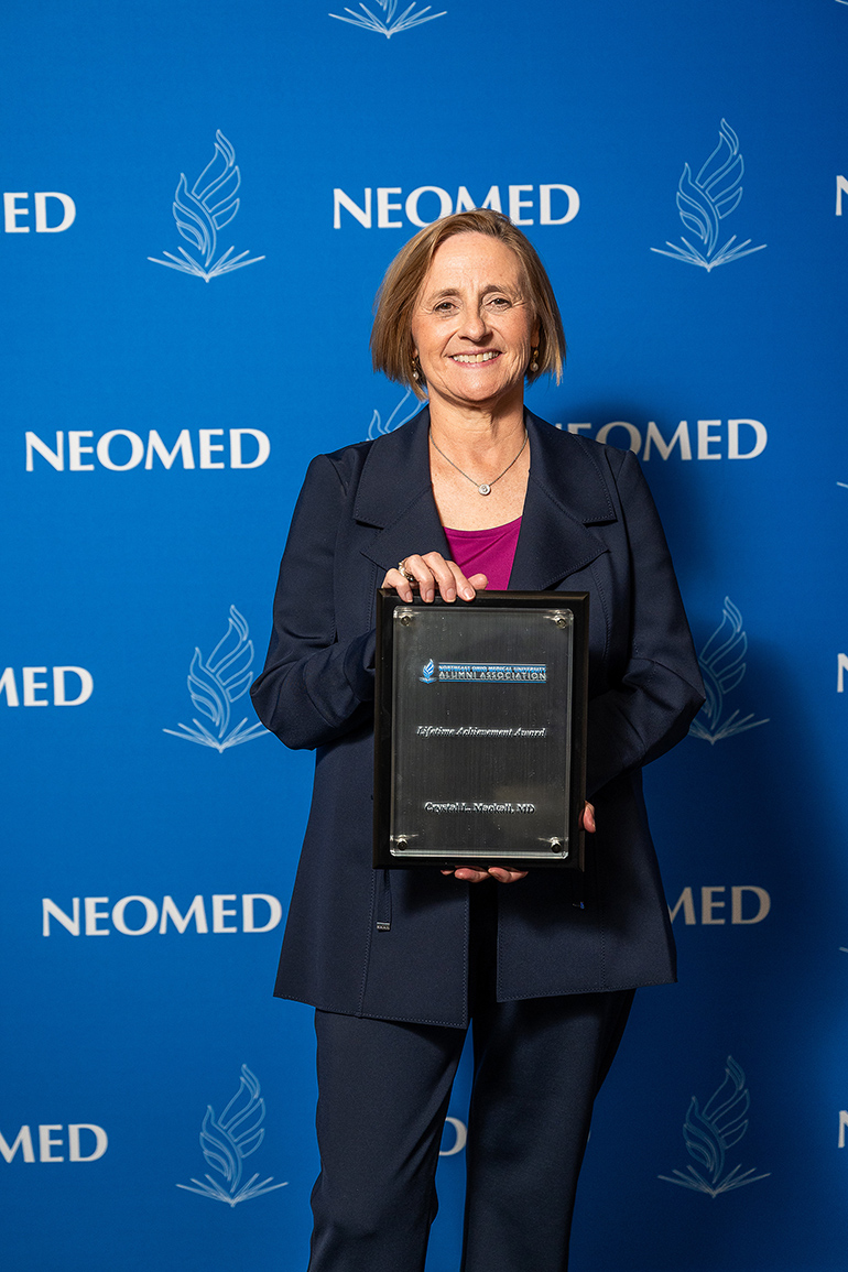 Crystal Mackall, in a suit, poses with an award plaque in front of a blue NEOMED backdrop
