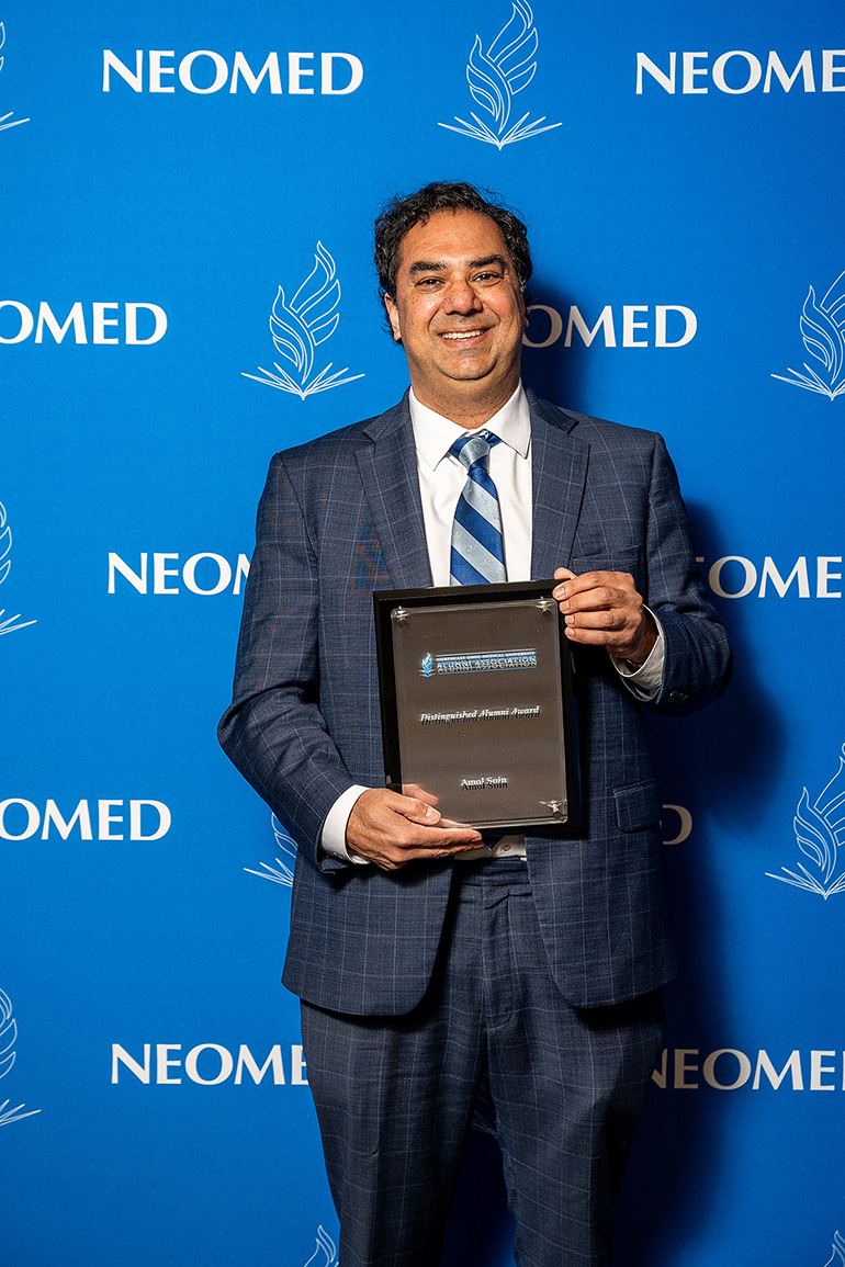 Amol Soin, in suit and tie, poses with an award plaque in front of a blue NEOMED backdrop