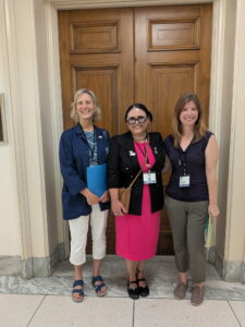 Three women in business casual attire pose outside a large wooden door