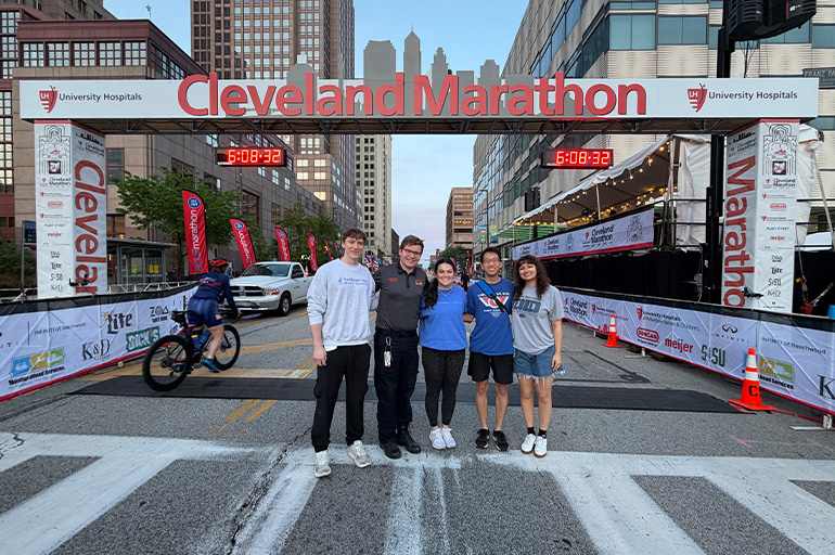 A group of five people in casual attire stand in a line, posing beneath a sign reading Cleveland Marathon