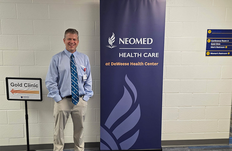 Man in blue shirt and tie poses next to a banner that says NEOMED Health Care at Deweese Health Center