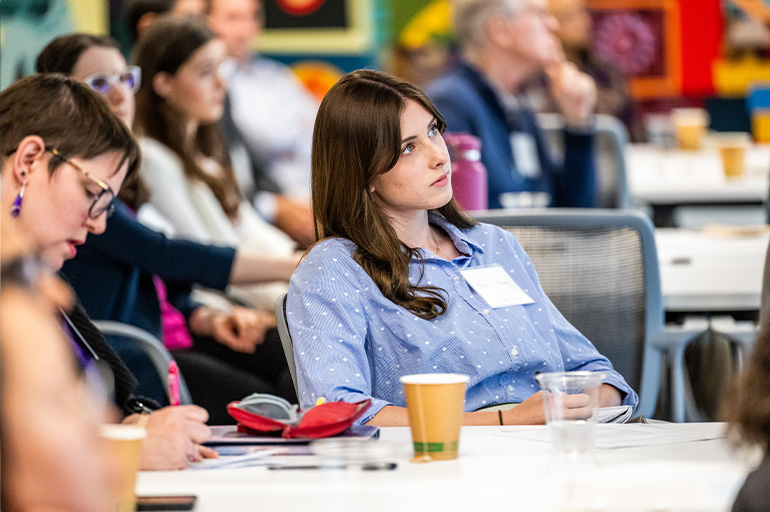 young woman in blue shirt sits at a table listening intently to an unseen speaker. A woman seated next to her takes notes