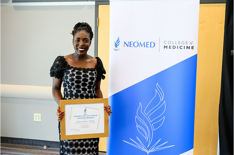 A smiling woman holding a framed certificate poses next to a banner for the NEOMED College of Medicine