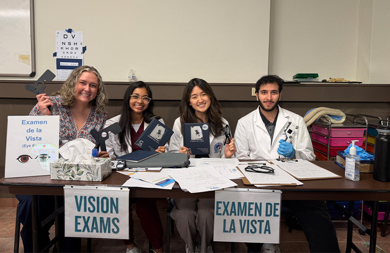 Four medical students in white coats sit at a table labeled "Vision Exams" and "Examen de la Vista"