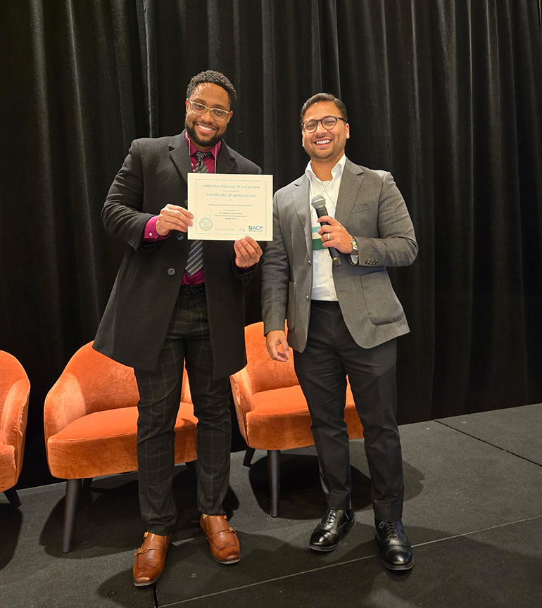 Two men wearing suits; one holds a certificate, the other holds a microphone