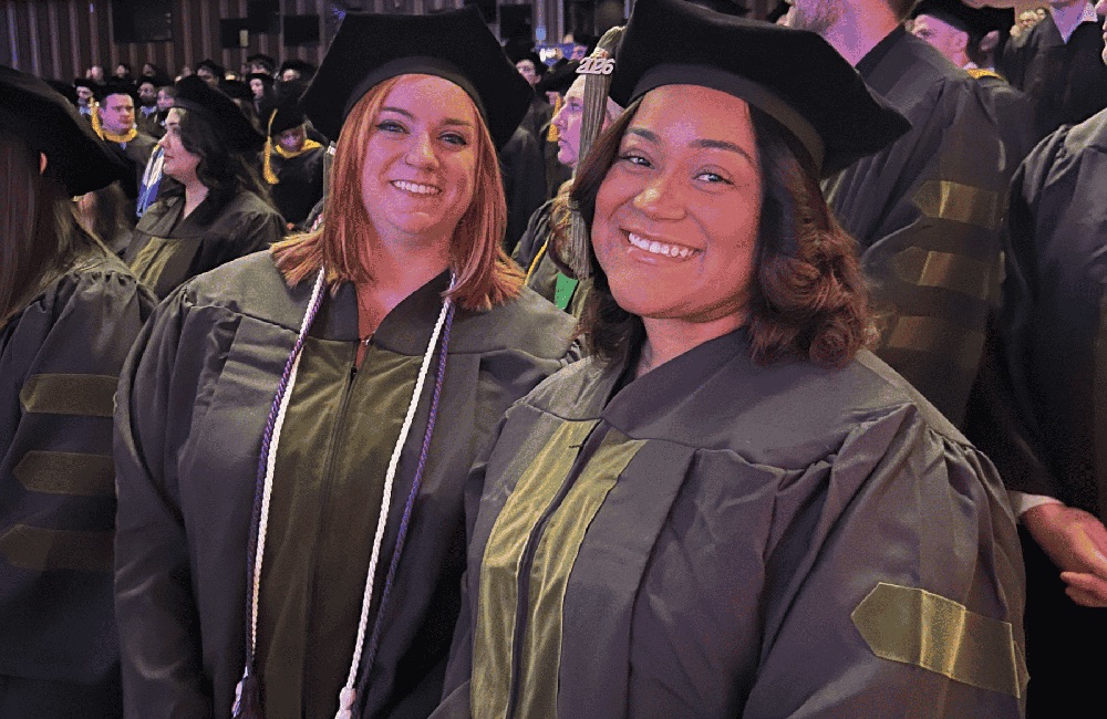 Two women graduates dressed in academic regalia smile at the camera