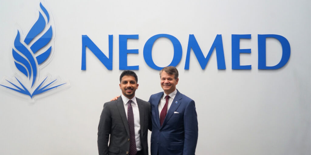 Two men wearing suits pose in front of the sign reading "NEOMED"