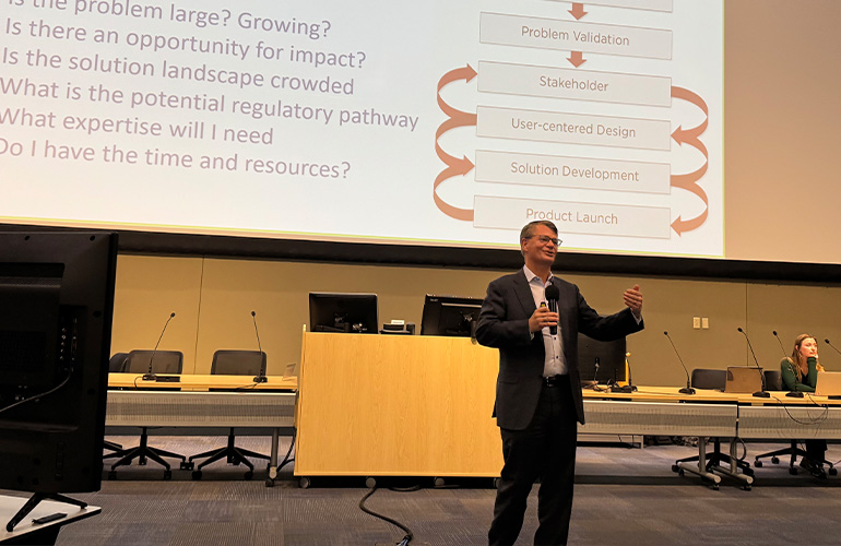 Man in dark suit presents in front of a Powerpoint presentation